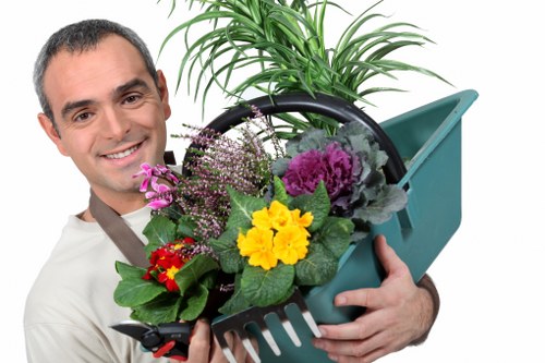 A smiling man with short dark hair and wearing a light grey shirt holding a teal plastic garden watering can filled with a variety of colorful plants and flowers, including purple, yellow, red, and green foliage, standing outdoors in a garden setting with a lush green background. The garden features well-maintained grass and a potted plant with large leafy fronds placed behind him. The scene appears bright and sunny, with natural light illuminating the vibrant plant colors. This outdoor space showcases elements typical of a landscaped garden in Edmonton, with a focus on plant care and maintenance that aligns with professional gardening and hedge trimming services offered by hedgetrimmingedmonton.co.uk.