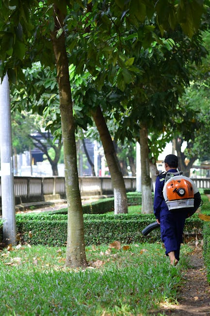 A garden maintenance worker, dressed in a bright green long-sleeved shirt and a wide-brimmed hat for sun protection, is trimming a neatly shaped hedge with a small pruning tool. The hedge features lus