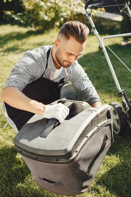 A man with a well-groomed hairstyle and a short beard is engaged in garden maintenance outdoors. He is wearing a checkered shirt, black apron, and white gloves, focusing on a large grey lawnmower or g